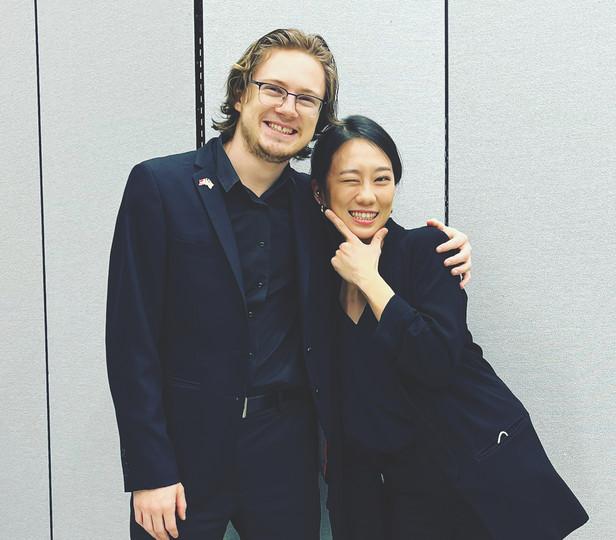 Two people, identified in the accompanying caption, stand side by side, smiling warmly. 两人都穿着黑色的衣服, 它们的背景是浅色的, 散发出友爱和喜悦的感觉.